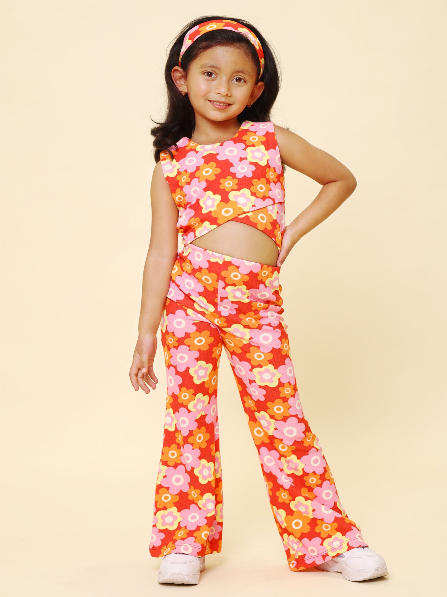 A young girl wearing a vibrant retro floral printed summer co-ord set, featuring a sleeveless cut-out top, flared pants, and a matching headband, posing with confidence against a pastel yellow background.