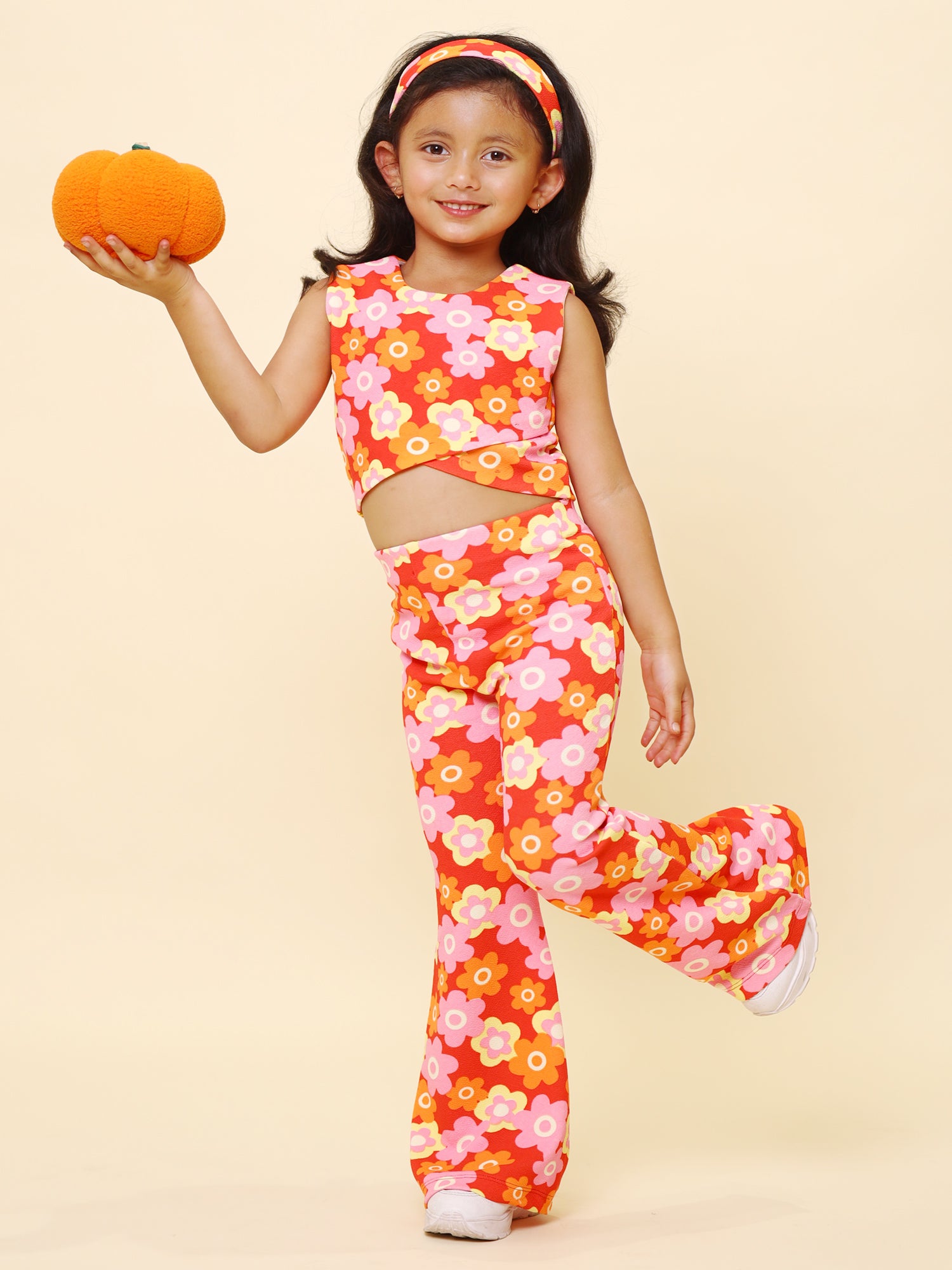 A young girl wearing a vibrant retro floral printed summer co-ord set, featuring a sleeveless cut-out top, flared pants, and a matching headband, posing with confidence against a pastel yellow background.