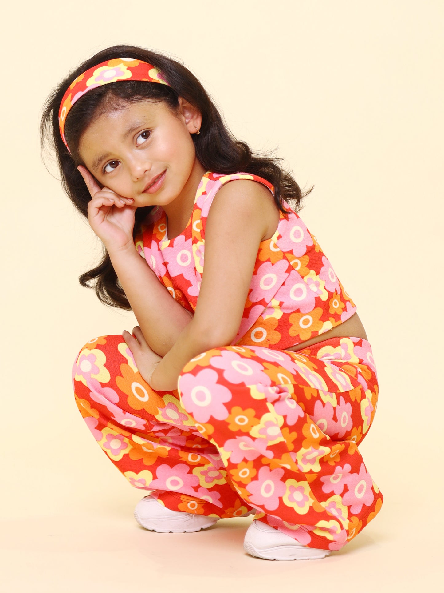 A young girl wearing a vibrant retro floral printed summer co-ord set, featuring a sleeveless cut-out top, flared pants, and a matching headband, posing with confidence against a pastel yellow background.