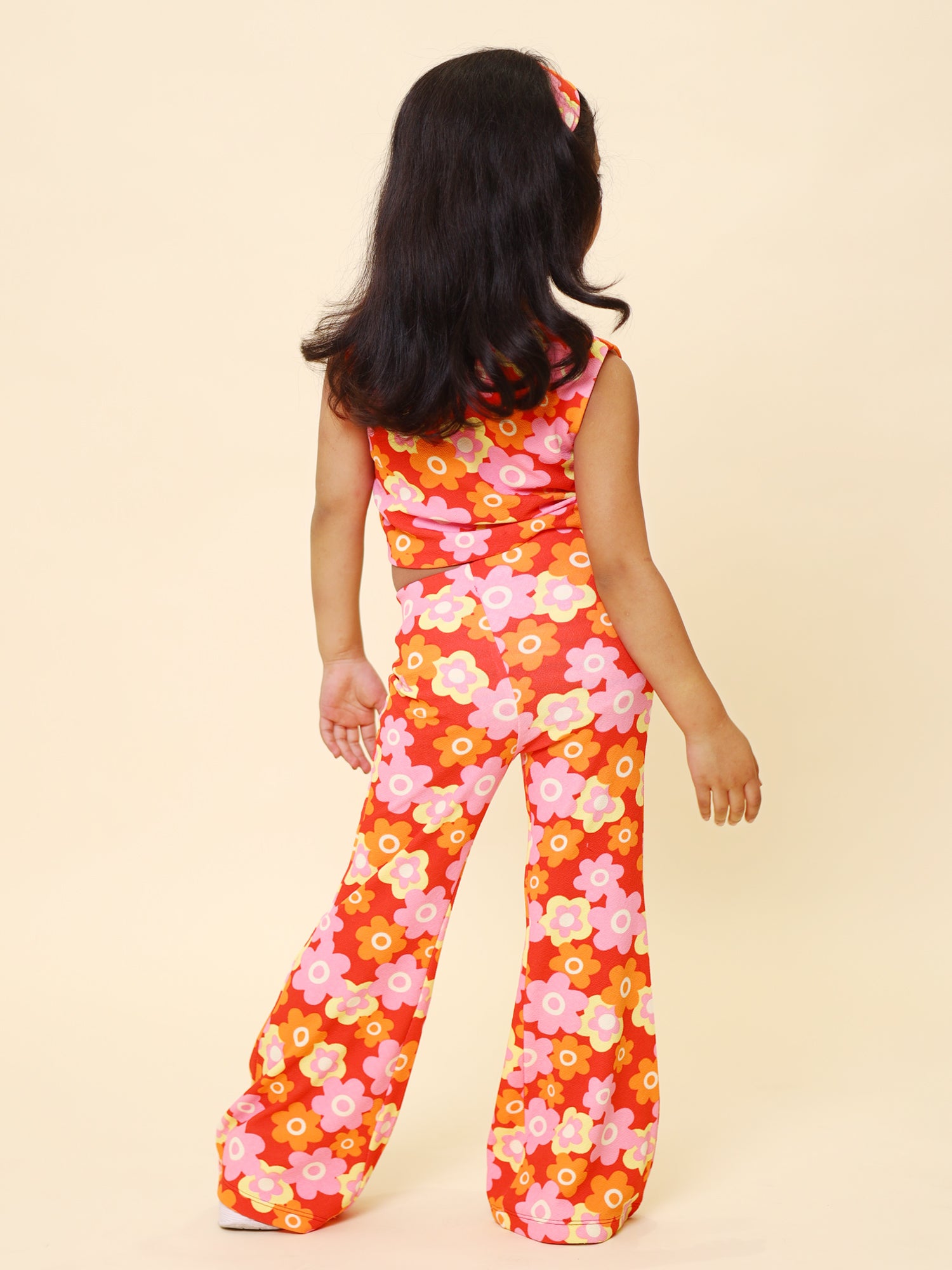 A young girl wearing a vibrant retro floral printed summer co-ord set, featuring a sleeveless cut-out top, flared pants, and a matching headband, posing with confidence against a pastel yellow background.