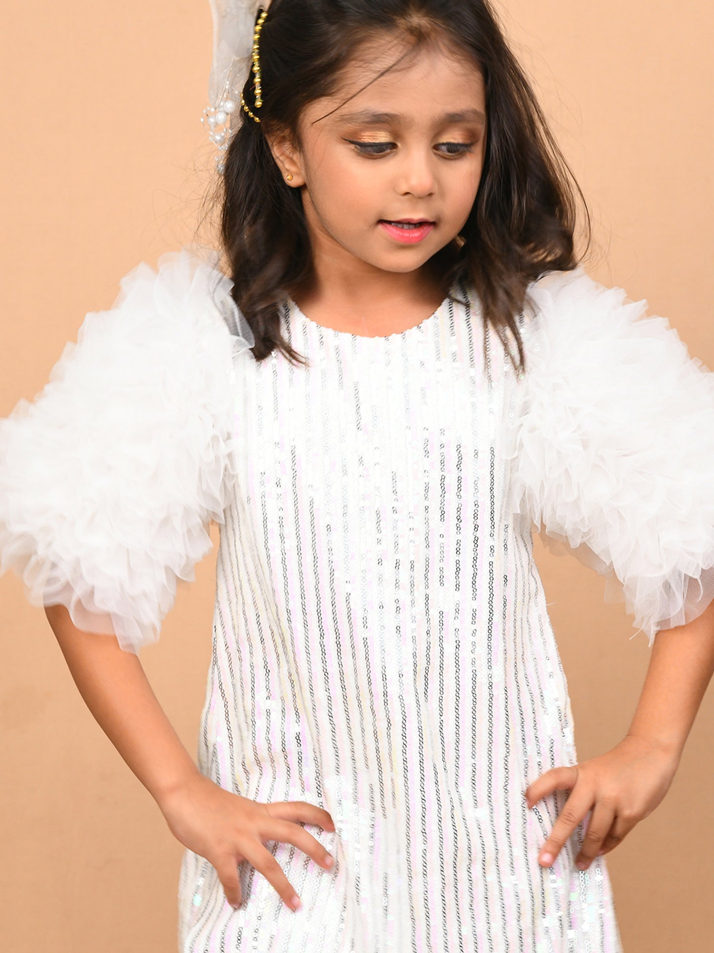 A young girl wearing a glamorous white sequin dress with voluminous tulle frill sleeves, paired with silver heels. The knee-length dress shimmers under the light, making it perfect for special occasions like parties and weddings. Ideal for B2B wholesale and white-label manufacturing by Sundaram Fashions.