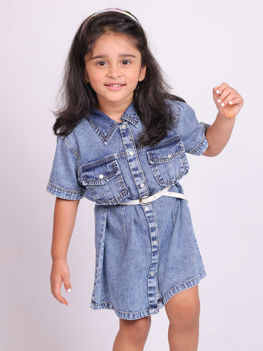 A young girl wearing a stylish denim shirt dress with short sleeves, snap buttons, and two chest pockets. The knee-length dress is cinched at the waist with a white belt, creating a structured yet comfortable fit. She pairs the look with trendy white and pink sneakers, posing confidently against a plain white background. This premium kidswear piece is ideal for casual outings and semi-formal occasions. Available for B2B wholesale and white-label manufacturing by Sundaram Fashions.